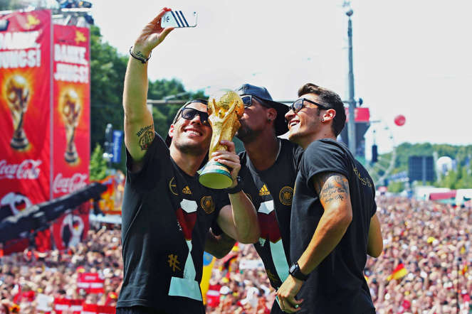 Germany's Lukas Podolski, Jerome Boateng and Mesut Oezil (L-R) pose for a 'selfie' with the World Cup trophy during celebrations to mark the team's 2014 Brazil World Cup victory, at a 'fan mile' public viewing zone in Berlin July 15, 2014.  Germany's victorious soccer team led by coach Joachim Loew returned home on Tuesday after winning the 2014 Brazil World Cup. A triumphant Germany team landed in Berlin on Tuesday to a hero's welcome, greeted by hundreds of thousands of jubilant Germans waving flags and wearing the national colours, revelling in the nation's fourth World Cup victory on Sunday in Brazil.