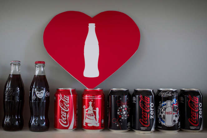 Bottles and cans of Coca-Cola beverages stand on display beneath; sign in an office at the Lanitis Bros Ltd. bottling plant, part of the Coca-Cola Hellenic Group, in Nicosia, Cyprus, on Tuesday, June 10, 2014.