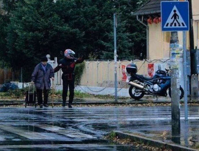 A motorcyclist pulls over to help an elderly lady cross the street on a rainy day.