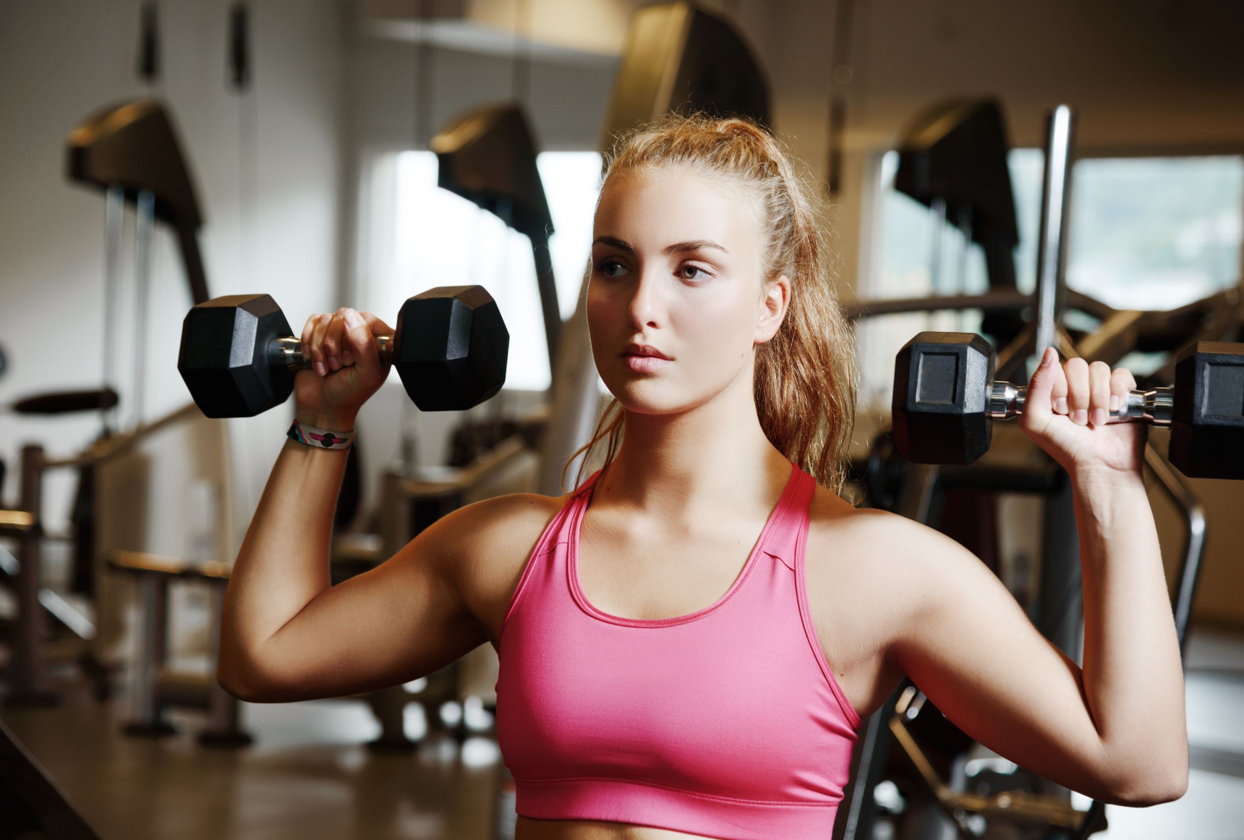 Beautiful woman wearing a pink top exercsing with dumbbells in a gym