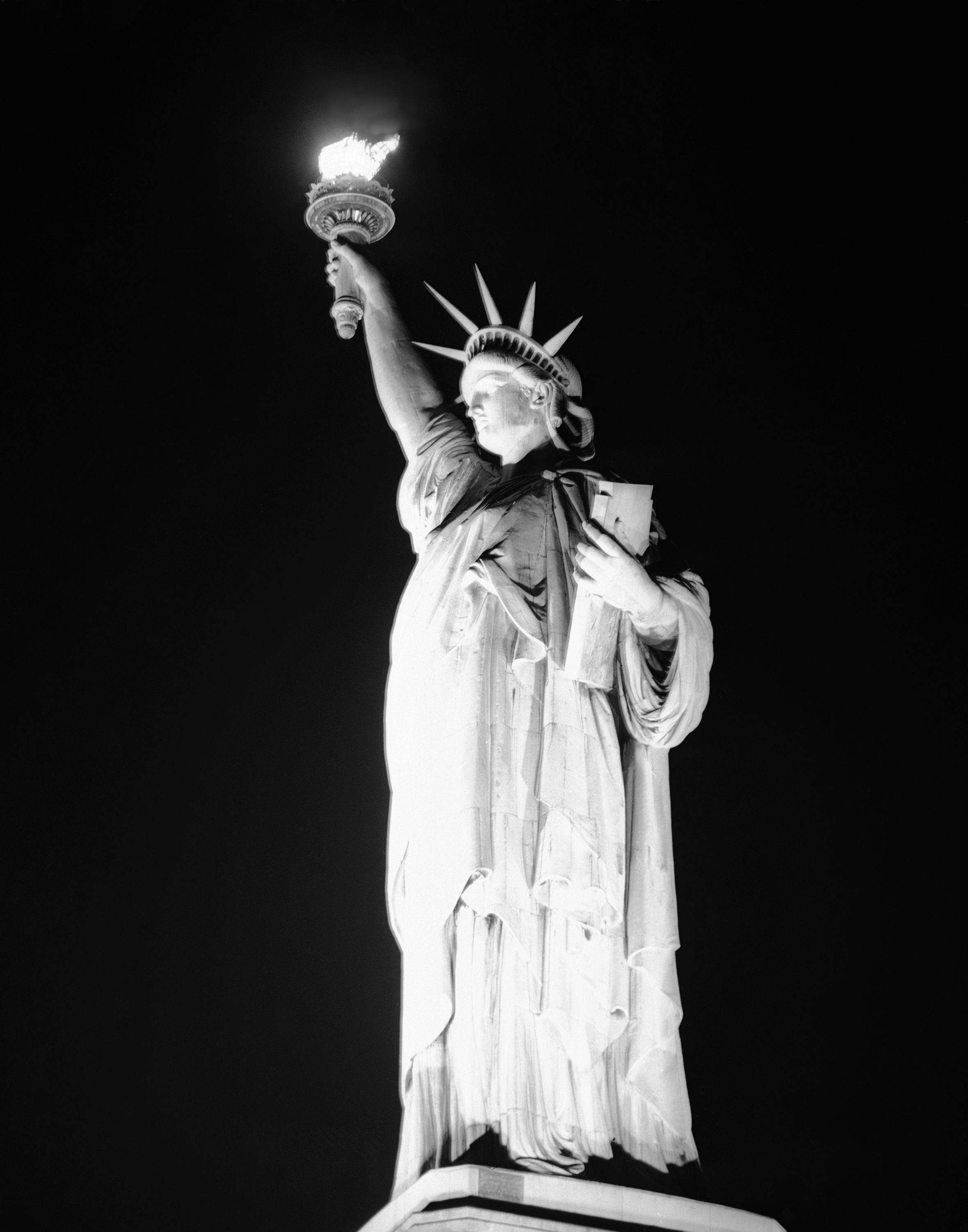 The torch of the Statue of Liberty blazes in the night as the lights are turned on once again at the island in New York Harbor, May 8, 1945, V-E Day, on which the official announcement of the unconditional surrender of Germany was proclaimed. Two service men stand guard at the base of the statue.